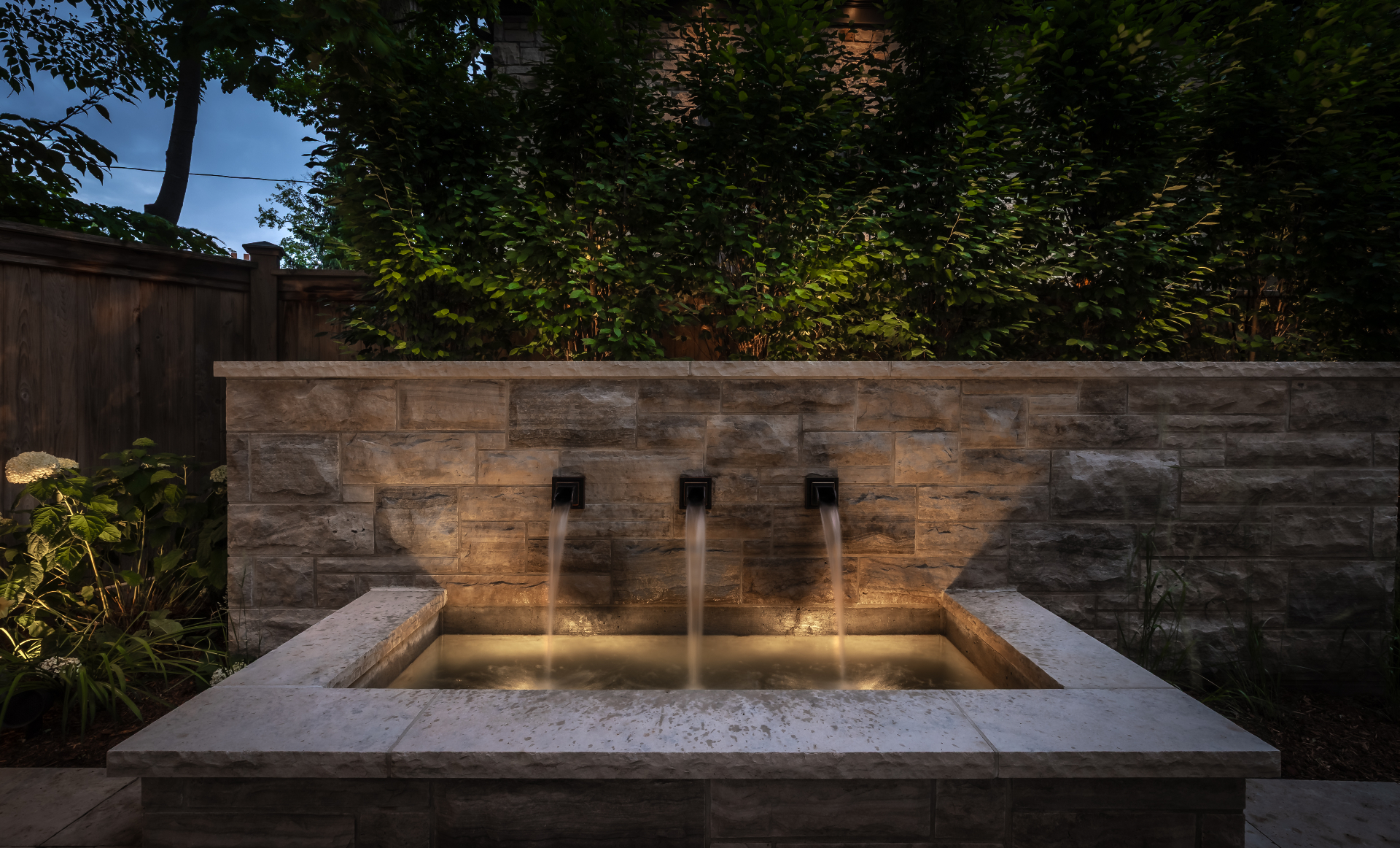 Illuminated triple-spout stone water wall at dusk